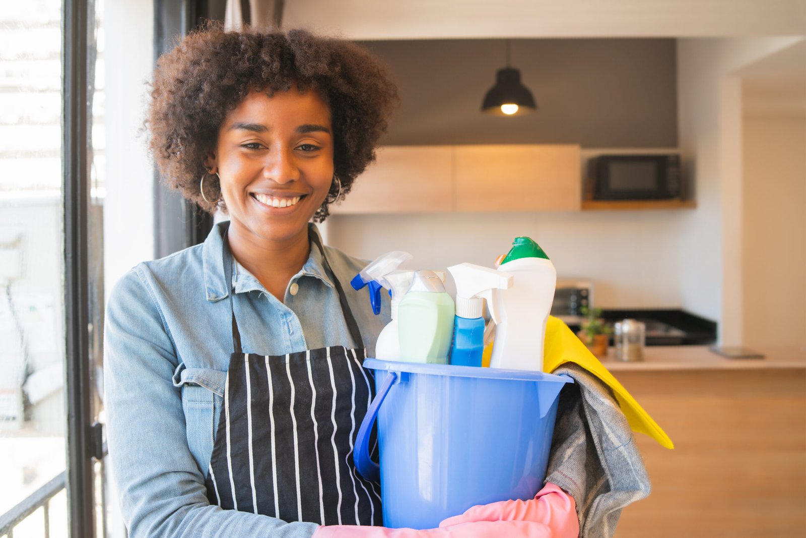 Portrait of young afro woman holding a bucket with cleaning items at home. Housekeeping and cleaning concept.